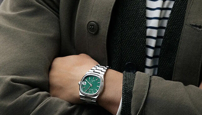 Man in casual blazer wearing Bulova stainless steel watch with green dial, arms crossed.